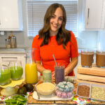 Hannah Magee wearing an orange dress and standing in her kitchen with two smoothies on the counter.