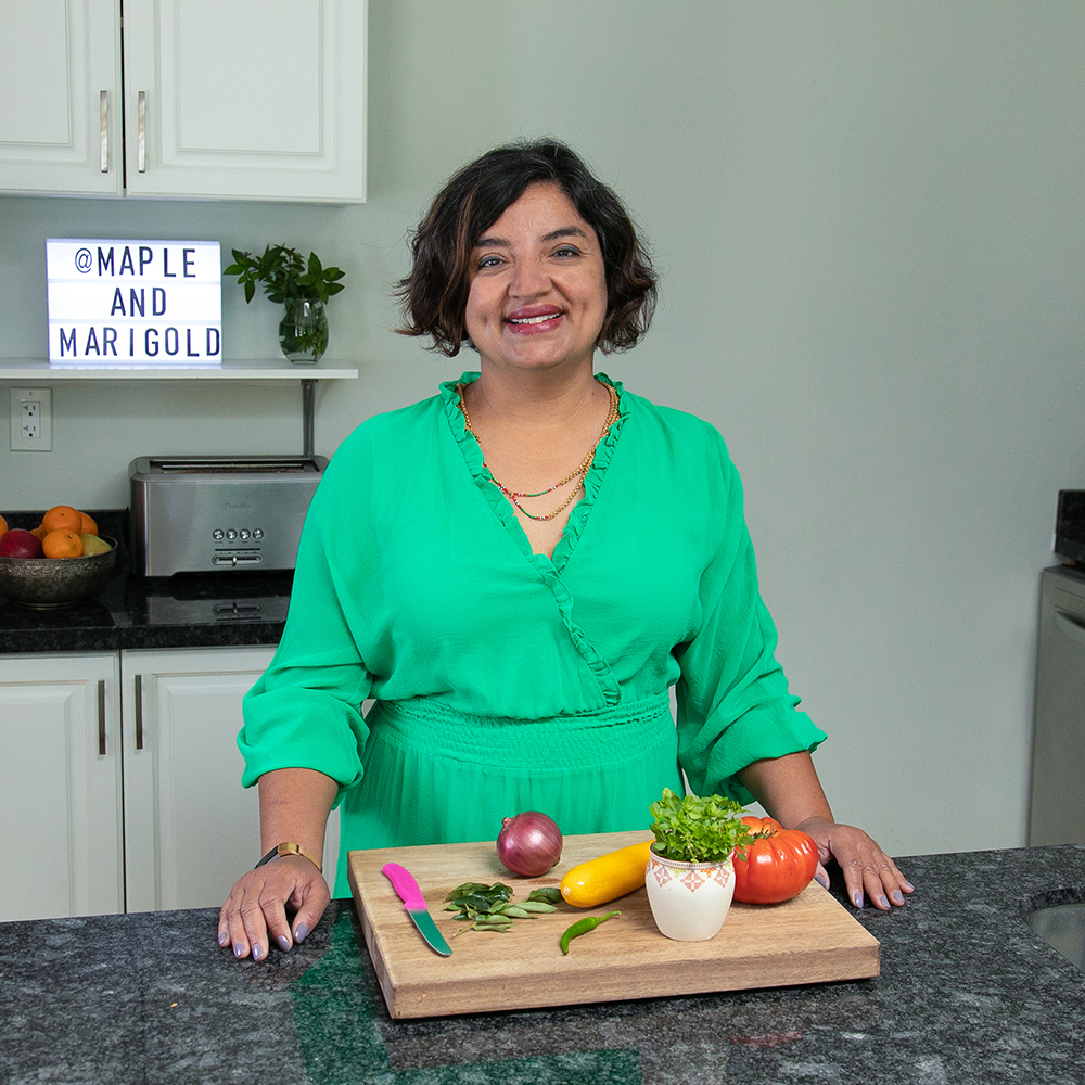 Puneeta wearing a green top and standing behind her kitchen counter with various food items on a cutting board.
