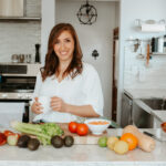 Tamara is wearing a white blouse, standing in her kitchen and holding a white mug. On the counter are oranges, avocados, tomatoes and celery.
